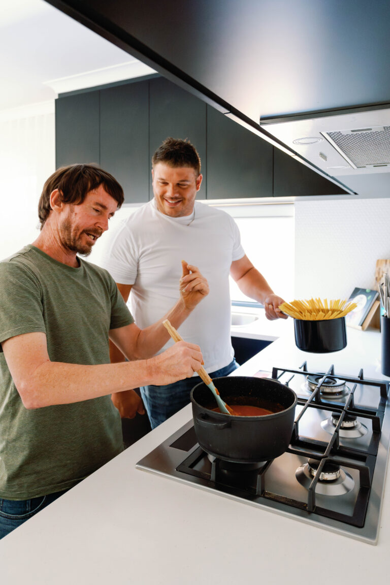 Two male housemates smiling whilst cooking pasta sauce and spaghetti over the stove