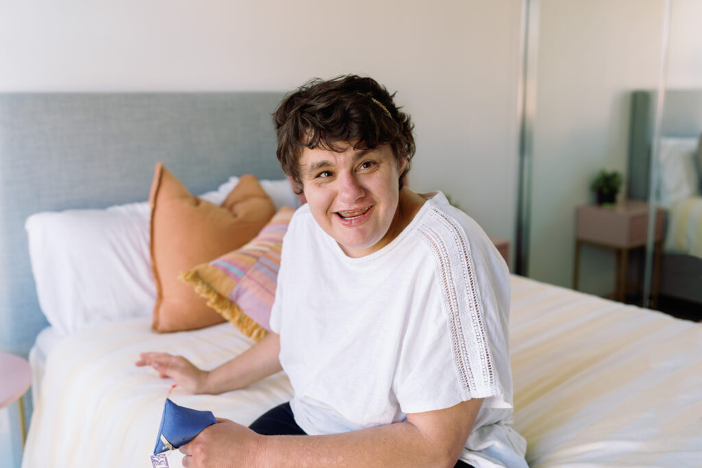 Girl in her 20s smiling, sitting on the edge of her bed